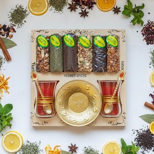 May include: A wooden tray with six spice packets, two red tea glasses, and a gold saucer. The tray is surrounded by a variety of herbs, spices, and citrus slices, including mint, star anise, and cinnamon sticks. The text "Yasmer Spices" is visible.