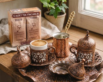 May include: A copper-toned coffee set featuring a round embossed tray, a small hammered pot with a long handle, and two white ceramic cups encased in intricate metal filigree holders with matching lids, alongside a box of coffee packets.