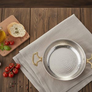 Puede incluir: Una sart&eacute;n de acero inoxidable con asas de lat&oacute;n descansa sobre un pa&ntilde;o de lino gris. A la izquierda hay una tabla de cortar de madera con aceite de oliva, pan y tomates. Un diente de ajo y m&aacute;s tomates tambi&eacute;n est&aacute;n sobre la mesa de madera.