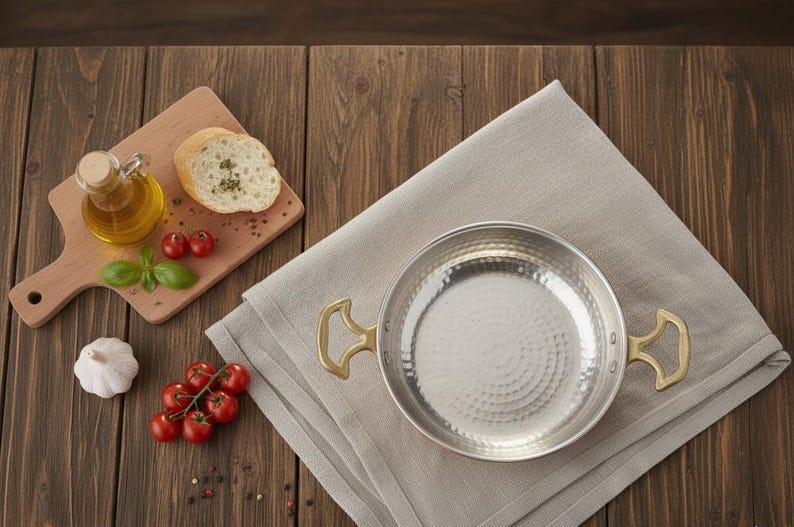 Puede incluir: Una sart&eacute;n de acero inoxidable con asas de lat&oacute;n descansa sobre un pa&ntilde;o de lino gris. A la izquierda hay una tabla de cortar de madera con aceite de oliva, pan y tomates. Un diente de ajo y m&aacute;s tomates tambi&eacute;n est&aacute;n sobre la mesa de madera.