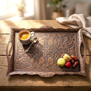 Pode incluir: Uma bandeja de servir retangular, cor de cobre, com intrincados padr&otilde;es florais e al&ccedil;as. A bandeja cont&eacute;m uma x&iacute;cara pequena de caf&eacute;, uma colher, macarons, framboesas e t&acirc;maras. A bandeja est&aacute; sobre uma superf&iacute;cie de madeira.