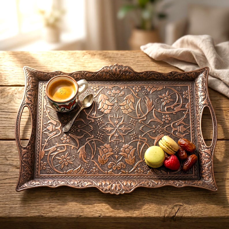 Pode incluir: Uma bandeja de servir retangular, cor de cobre, com intrincados padr&otilde;es florais e al&ccedil;as. A bandeja cont&eacute;m uma x&iacute;cara pequena de caf&eacute;, uma colher, macarons, framboesas e t&acirc;maras. A bandeja est&aacute; sobre uma superf&iacute;cie de madeira.