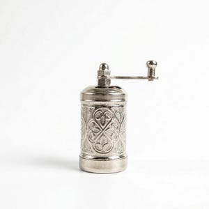 Puede incluir: Un molinillo de especias de metal plateado con un intrincado patr&oacute;n floral en relieve en su cuerpo cil&iacute;ndrico. La parte superior del molinillo incluye una peque&ntilde;a tuerca y un mango horizontal con un pomo redondeado.