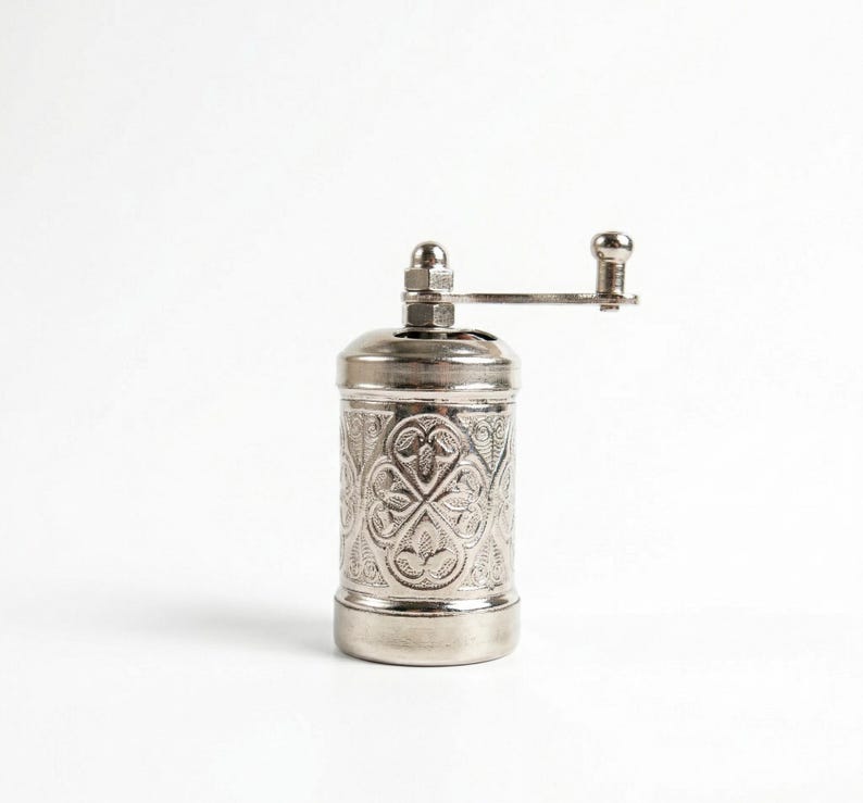 Puede incluir: Un molinillo de especias de metal plateado con un intrincado patr&oacute;n floral en relieve en su cuerpo cil&iacute;ndrico. La parte superior del molinillo incluye una peque&ntilde;a tuerca y un mango horizontal con un pomo redondeado.