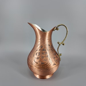 Puede incluir: Una jarra de color cobre con un dise&ntilde;o ornamentado grabado en la superficie. La jarra tiene un interior plateado y un asa de lat&oacute;n. El dise&ntilde;o presenta un patr&oacute;n repetitivo de formas estilizadas. La jarra probablemente se utiliza para servir bebidas.
