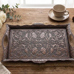Pode incluir: Uma bandeja de metal retangular ornamentada com al&ccedil;as, apresentando um padr&atilde;o floral e de folhas detalhado. A bandeja &eacute; de cor prata antiga escura e est&aacute; sobre uma superf&iacute;cie de madeira, junto com uma x&iacute;cara de ch&aacute; e livros.