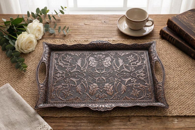 Pode incluir: Uma bandeja de metal retangular ornamentada com al&ccedil;as, apresentando um padr&atilde;o floral e de folhas detalhado. A bandeja &eacute; de cor prata antiga escura e est&aacute; sobre uma superf&iacute;cie de madeira, junto com uma x&iacute;cara de ch&aacute; e livros.