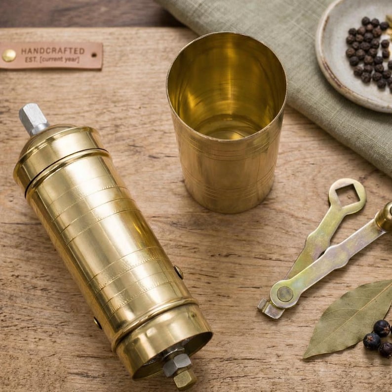 May include: A handcrafted brass spice grinder and a matching brass cup sit on a wooden surface. Two small wrenches, a bay leaf, and peppercorns are also present. The grinder has a cylindrical shape with a textured surface.