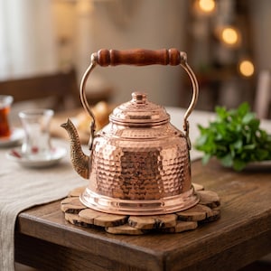 May include: A copper tea kettle with a hammered texture, a wooden handle, and a decorative spout. The kettle sits on a wooden coaster. The design is traditional and the colour is a warm copper.