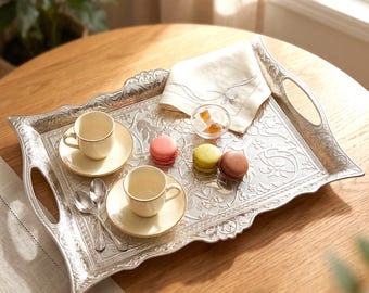 May include: A silver serving tray with ornate floral detailing holds two small teacups with saucers, two spoons, a folded napkin, a small bowl of sugar cubes, and three colorful macarons. The tray has handles and is set on a wooden table.