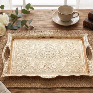 Pode incluir: Uma bandeja de servir dourada decorativa com um padr&atilde;o floral ornamentado. A bandeja tem al&ccedil;as e est&aacute; sobre um tapete tecido. Uma ch&aacute;vena e pires, rosas e livros tamb&eacute;m est&atilde;o na mesa.