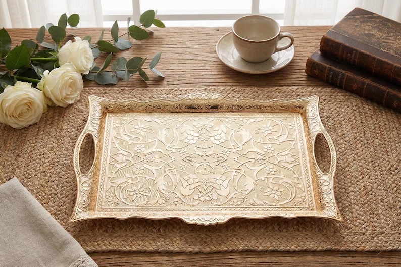 Pode incluir: Uma bandeja de servir dourada decorativa com um padr&atilde;o floral ornamentado. A bandeja tem al&ccedil;as e est&aacute; sobre um tapete tecido. Uma ch&aacute;vena e pires, rosas e livros tamb&eacute;m est&atilde;o na mesa.
