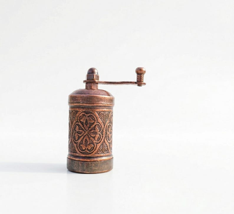 Puede incluir: Un molinillo de especias cil&iacute;ndrico de color cobre con un intrincado patr&oacute;n floral en relieve en el cuerpo. El dispositivo incluye una manivela manual montada en la parte superior con un peque&ntilde;o pomo, dise&ntilde;ada para moler especias girando.