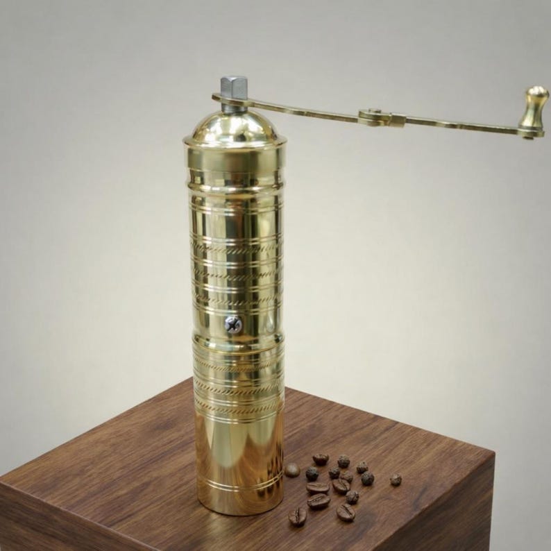 May include: A gold-colored manual coffee grinder with a cylindrical body and a crank handle. The grinder is made of metal and has decorative horizontal lines. Coffee beans are scattered on the wooden surface next to the grinder.