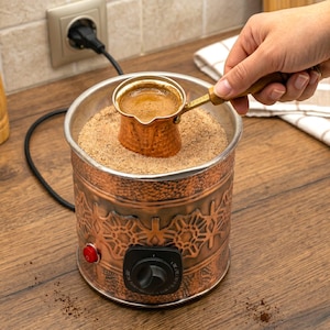 Puede incluir: Una cafetera eléctrica de cobre con una pequeña cafetera de cobre en su interior. La cafetera está llena de café. La cafetera tiene un diseño decorativo y un botón rojo de encendido. La cafetera está enchufada.