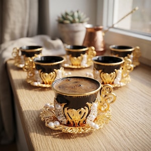 Könnte beinhalten: Sechs schwarze Kaffeetassen mit goldenen Filigran-Griffen und Perlenakzenten. Die Tassen sind mit Kaffee gefüllt. Eine Kupfer-Kaffeekanne und eine Sukkulente befinden sich im Hintergrund, auf einer Holzoberfläche.