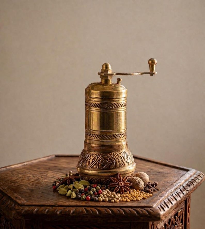 May include: A vintage brass pepper mill with a hand crank, standing on a carved wooden surface. The mill is surrounded by a variety of whole spices, including peppercorns, star anise, and cardamom. The warm tones of the brass and wood create a rustic aesthetic.