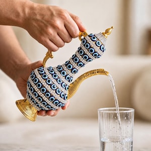 May include: A decorative gold-coloured pitcher with a blue and white design, pouring water into a clear glass. The pitcher is adorned with blue eye-shaped embellishments and clear crystals. The pitcher has a height of approximately 25 cm.