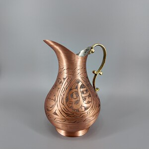 Puede incluir: Una jarra de color cobre con un dise&ntilde;o grabado y ornamentado. La jarra tiene un borde plateado y un asa dorada. El cuerpo de la jarra presenta un patr&oacute;n detallado de l&iacute;neas y formas arremolinadas, lo que aumenta su atractivo decorativo.