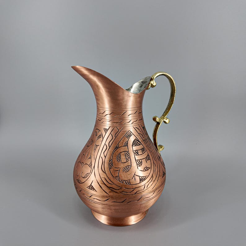 Puede incluir: Una jarra de color cobre con un dise&ntilde;o grabado y ornamentado. La jarra tiene un borde plateado y un asa dorada. El cuerpo de la jarra presenta un patr&oacute;n detallado de l&iacute;neas y formas arremolinadas, lo que aumenta su atractivo decorativo.