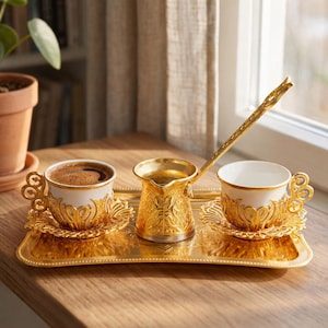 Könnte beinhalten: Ein goldfarbenes Kaffeeservice auf einem rechteckigen Tablett. Das Set umfasst eine kleine Kaffeekanne, zwei Tassen und Untertassen, alle mit aufwendigen Designs. Die Tassen sind weiß mit goldenen Verzierungen, und eine ist mit Kaffee gefüllt. Ein Löffel mit langem Griff liegt auf dem Tablett.