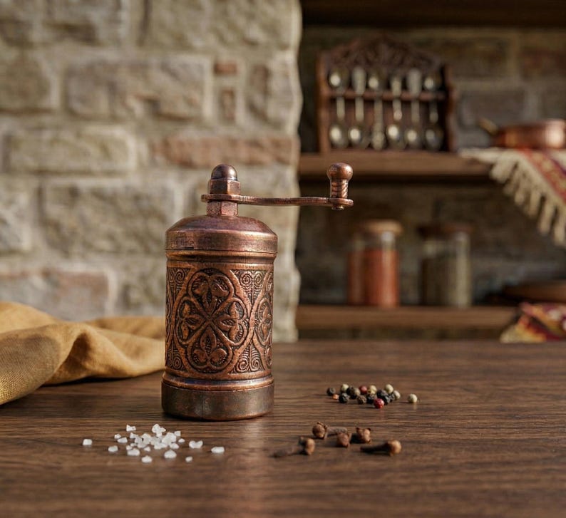Puede incluir: Un molinillo de especias de metal en tono cobre con un intrincado patr&oacute;n floral en relieve en el cuerpo cil&iacute;ndrico y una manivela manual en la parte superior. El molinillo est&aacute; junto a peque&ntilde;os montones de sal blanca y granos de pimienta.