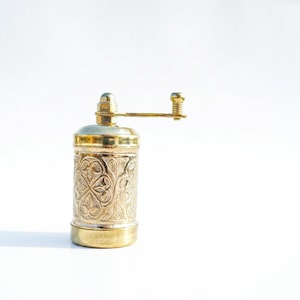 Puede incluir: Un molinillo de especias cil&iacute;ndrico de lat&oacute;n con un intrincado patr&oacute;n floral en relieve en el cuerpo. El molinillo incluye un mango horizontal montado en la parte superior con un peque&ntilde;o pomo, una base lisa y un acabado met&aacute;lico pulido.