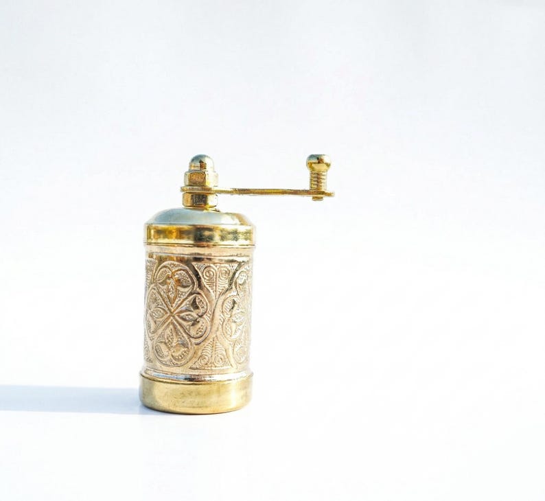 Puede incluir: Un molinillo de especias cil&iacute;ndrico de lat&oacute;n con un intrincado patr&oacute;n floral en relieve en el cuerpo. El molinillo incluye un mango horizontal montado en la parte superior con un peque&ntilde;o pomo, una base lisa y un acabado met&aacute;lico pulido.