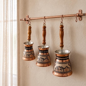 May include: Three copper coffee pots with wooden handles hanging from a copper rail. Each pot features a flared rim, a tapered body, and a dark decorative band with intricate floral patterns. The pots vary in size, arranged from smallest to largest.