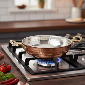 Puede incluir: Una sart&eacute;n de color cobre con interior martillado y asas de lat&oacute;n se encuentra sobre una estufa de gas con una llama azul. El entorno de la cocina incluye tomates frescos, ajo y aceite de oliva.