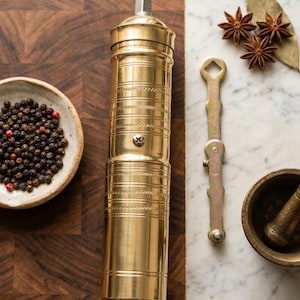 May include: A gold-colored pepper mill with a textured design, alongside a small bowl of peppercorns, star anise, and a mortar and pestle. The items are arranged on a wooden cutting board and a marble surface.