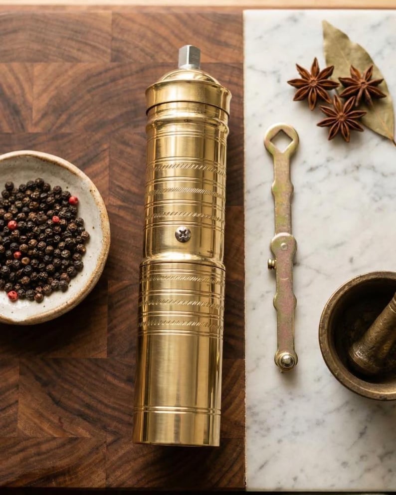 May include: A gold-colored pepper mill with a textured design, alongside a small bowl of peppercorns, star anise, and a mortar and pestle. The items are arranged on a wooden cutting board and a marble surface.