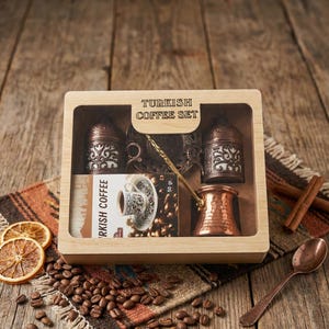 Può includere: Un set in scatola di legno con l'etichetta "TURKISH COFFEE SET" contiene una caffettiera in rame, due contenitori decorativi, un cucchiaino e una carta con un disegno di tazza di caffè. Il set è esposto su un tessuto fantasia con chicchi di caffè, fette d'arancia essiccate e bastoncini di cannella.