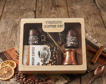 May include: A Turkish coffee set in a wooden box, including a copper coffee pot, two decorative containers, a small spoon, and a card with a coffee cup design. The box is on a patterned textile with coffee beans and dried orange slices.
