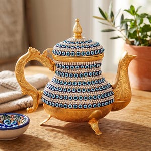 May include: A gold-toned metal teapot featuring an ornate handle and spout, with a body and lid covered in rows of blue and white evil eye beads and clear rhinestones. The teapot stands on three decorative feet and has a pointed finial on the lid.