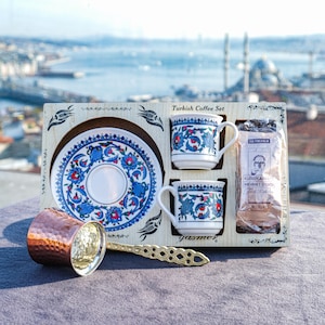 Puede incluir: Un juego de café turco presentado en una caja de madera. Incluye dos tazas pequeñas, un platillo y una bolsa de café. Las tazas y el platillo presentan un diseño floral azul y rojo. También se incluye una cafetera de cobre y una cuchara decorativa.