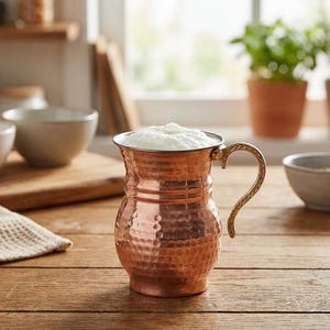 May include: A copper pitcher with a hammered finish and an ornate handle, filled with a white, foamy liquid. The pitcher is on a wooden table. Kitchenware is the theme.
