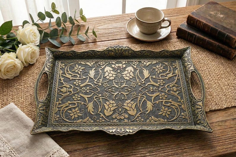 Pode incluir: Uma bandeja retangular em estilo antigo com intrincados padr&otilde;es florais e de folhas douradas sobre um fundo escuro. A bandeja tem al&ccedil;as em cada lado e uma borda decorativa. Uma x&iacute;cara e um pires, livros e flores est&atilde;o por perto.