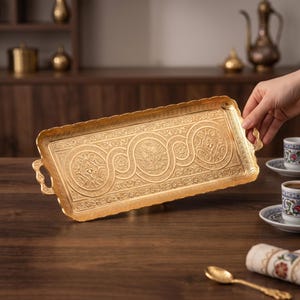 Pode incluir: Uma bandeja de servir retangular de metal na cor dourada com padrões em relevo intrincados, bordas recortadas e duas alças decorativas nas laterais. A superfície exibe motivos florais e geométricos detalhados em toda a base.