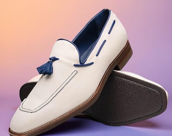 May include: White suede loafers with navy blue accents, including a tassel and interior lining. The shoes have a brown sole and stitching details. The loafers are a stylish footwear option.