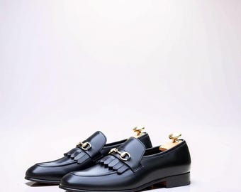 Pode incluir: Um par de mocassins de couro preto com um detalhe de bridão em metal dourado no peito do pé e uma aba com franjas. Cada sapato inclui uma forma de madeira com um botão dourado inserido na zona do calcanhar.