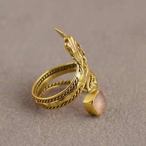 Peut inclure: Une bague dor&eacute;e au design orn&eacute;. La bague pr&eacute;sente un anneau enroul&eacute; avec un motif textur&eacute; et un &eacute;l&eacute;ment d&eacute;coratif qui s'&eacute;tend vers le haut. Une pierre pr&eacute;cieuse rose clair carr&eacute;e est suspendue &agrave; la bague.