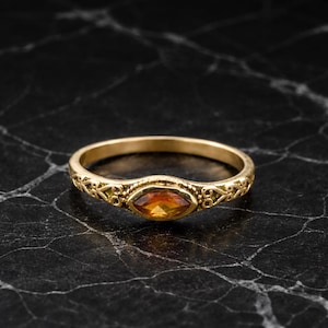 May include: A gold ring with an oval, orange gemstone. The ring has an ornate, filigree design on the band. The gemstone is set in a bezel setting. The ring is displayed on a black marble surface.