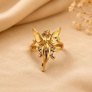 May include: A gold-toned ring featuring a central figure of a winged being with detailed, textured wings and a slender form. The metal has a polished, reflective finish, and the band is integrated into the design, extending from the figure's sides.