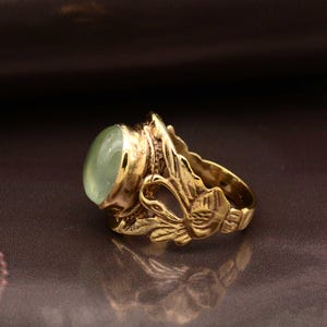 Peut inclure: Une bague dorée ornée d'une pierre ovale vert clair. La bague présente un design orné avec des détails de feuilles et de fleurs. La bague est posée sur un fond marron foncé, soulignant le design et la couleur complexes de la bague.
