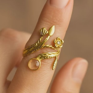 May include: A gold-colored ring with a floral and leaf design. The ring features a small flower with a central gem, a light-colored gemstone, and delicate leaf and stem details. The ring is worn on a finger.