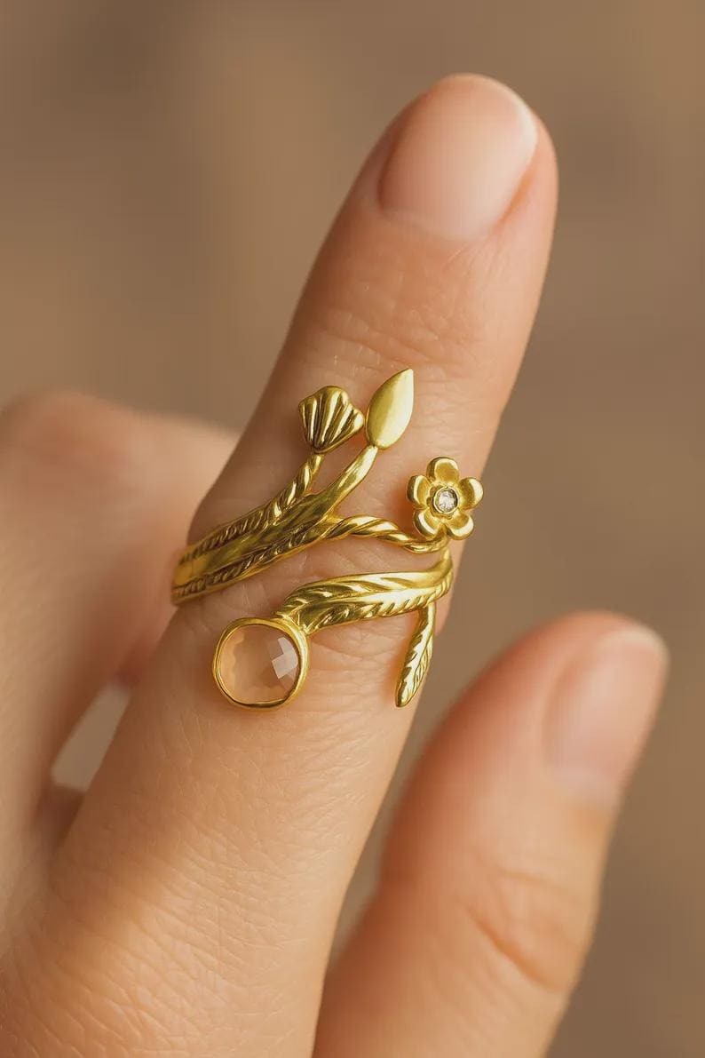 Peut inclure: Une bague dor&eacute;e avec un motif floral et de feuilles. La bague pr&eacute;sente une petite fleur avec un bijou central, une pierre pr&eacute;cieuse de couleur claire et des d&eacute;tails d&eacute;licats de feuilles et de tiges. La bague est port&eacute;e sur un doigt.