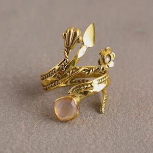 Peut inclure: Une bague dor&eacute;e au design unique. La bague est orn&eacute;e d'une pierre pr&eacute;cieuse rose p&acirc;le et de d&eacute;tails floraux et en forme de coquillage. La bague s'enroule autour du doigt et a une apparence d&eacute;corative et artistique.