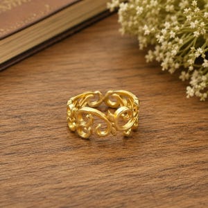 May include: A gold-toned ring featuring an intricate openwork design composed of repeating swirling scroll patterns and leaf-like motifs. The band has a polished metallic finish and a continuous, interconnected structure that creates a delicate, airy appearance.