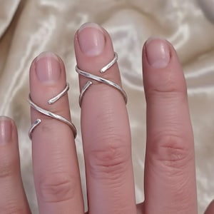 May include: Close-up of three silver rings worn on fingers. The rings have a minimalist design, with a twisted or wrapped appearance. The rings are made of a shiny, metallic material, and they are set against a soft, neutral background.